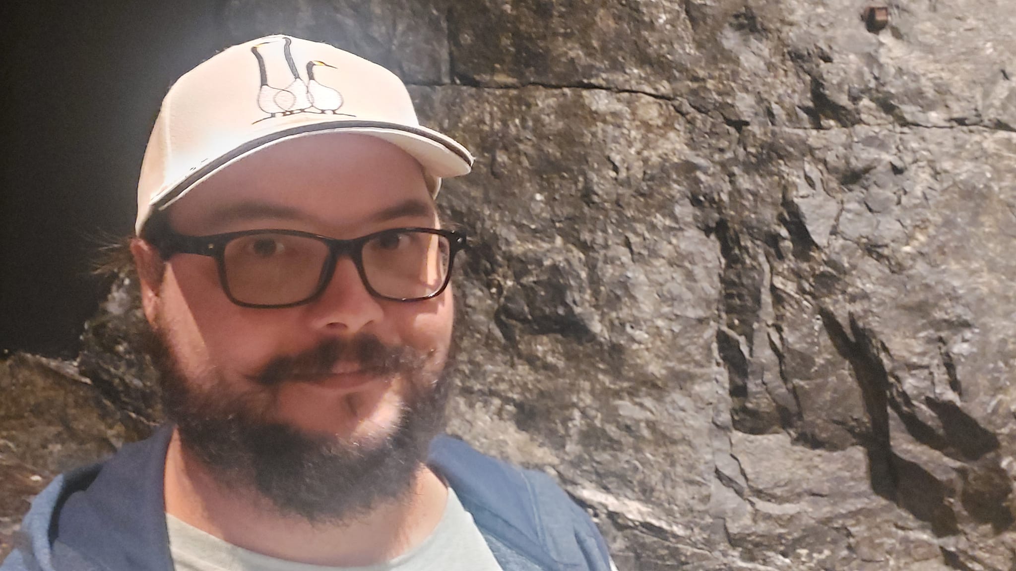 A man with a ball cap and bushy facial hair is standing in front of a hard rock tunnel looking contented